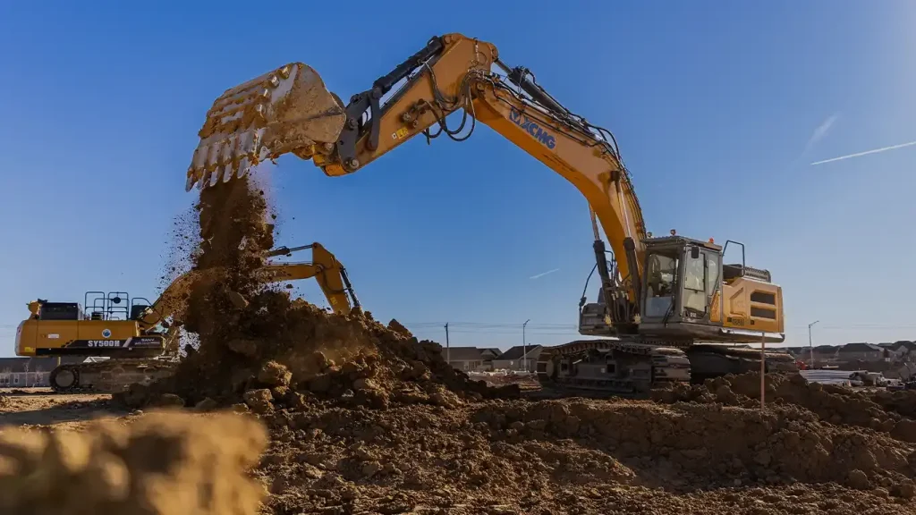 A ReadyDig XCMG excavator in action on a construction site, lifting and moving dirt with a large bucket under a clear blue sky.